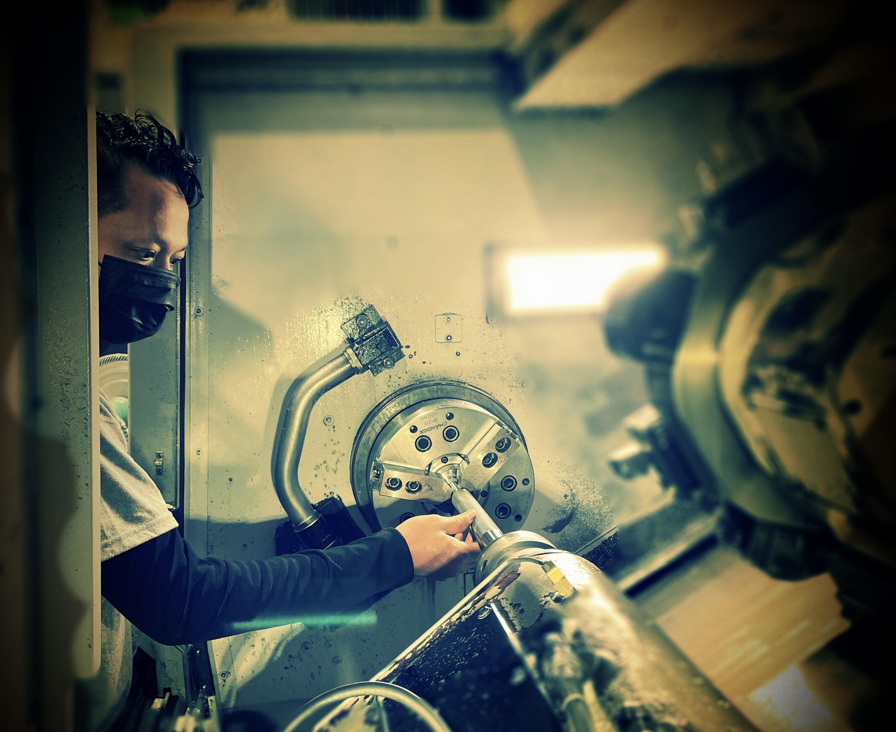 A side-view shot of a machinist, wearing a black mask and a white t-shirt with dark long sleeves, working at a large CNC lathe machine.