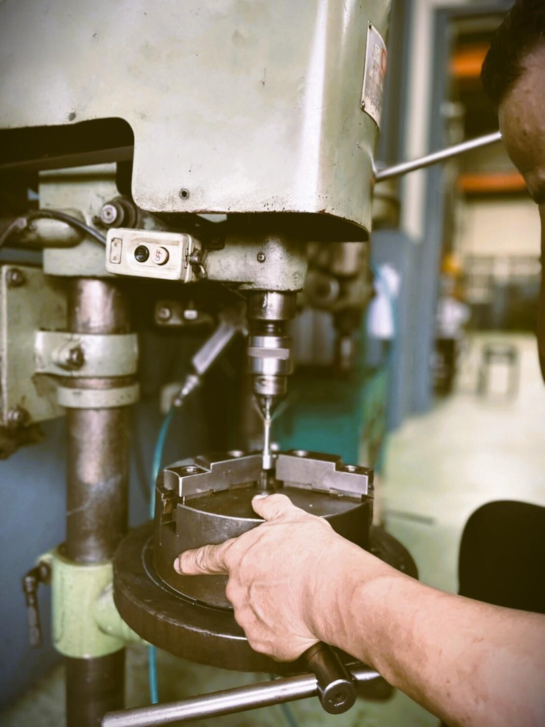 A close-up side view of an industrial drill press in action. A worker, with their hand resting on a chuck, is operating the machine as a drill bit touches a metal workpiece. 